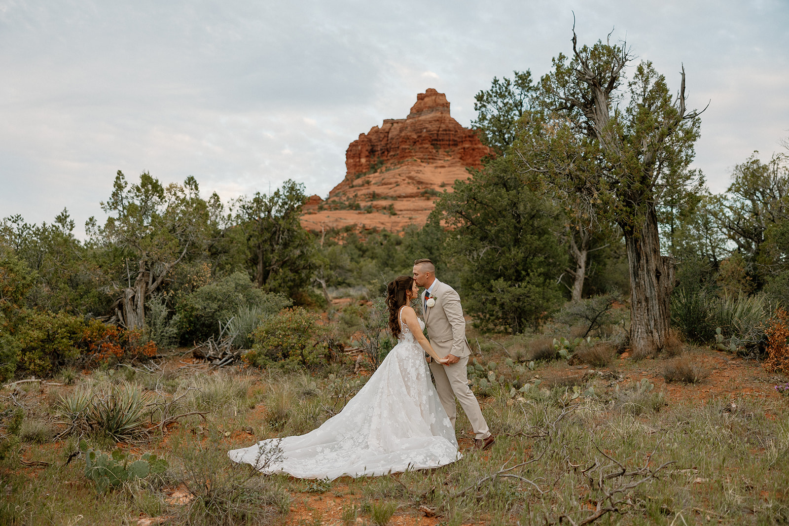 The couple shares a kiss in a quiet patch of desert greenery, her long train pooling in the grass with Bell Rock peeking through the trees in the background.