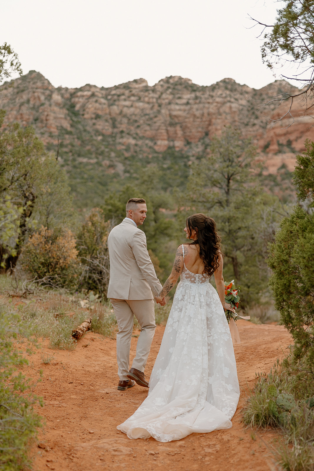 Groom glances over his shoulder at the bride as they walk together through a wooded Sedona trail, her bouquet of warm florals tucked at her side.