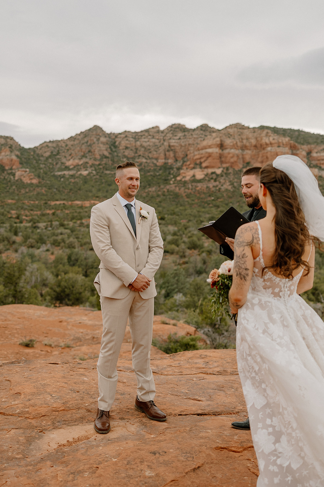 Bride joins the groom and officiant on a red rock platform with Bell Rock Sedona and pine-covered cliffs framing their elopement ceremony.