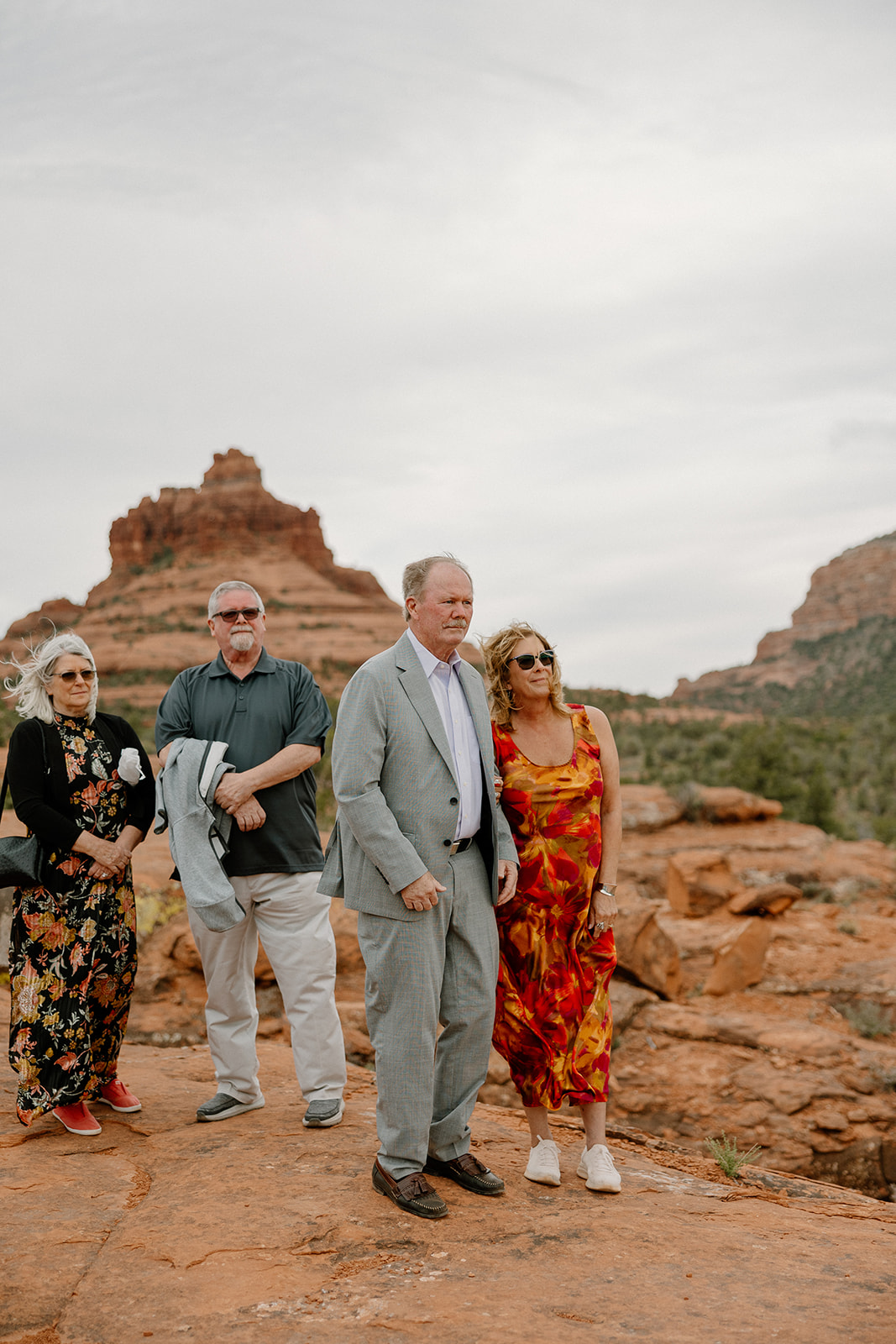 A small group of guests stands on red rock ledges, gazing toward the ceremony with Bell Rock Sedona rising dramatically behind them.