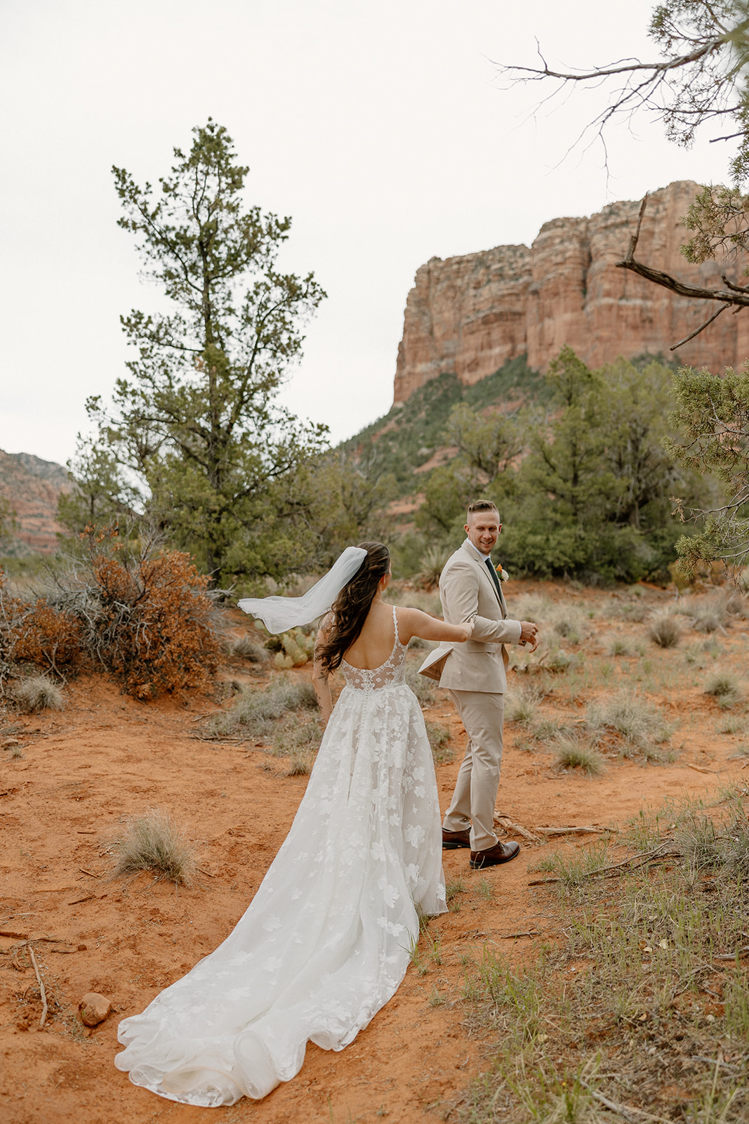 Groom glances back playfully at his bride, who follows him along the red dirt trail in her flowing lace gown, with Bell Rock Sedona rising in the background.