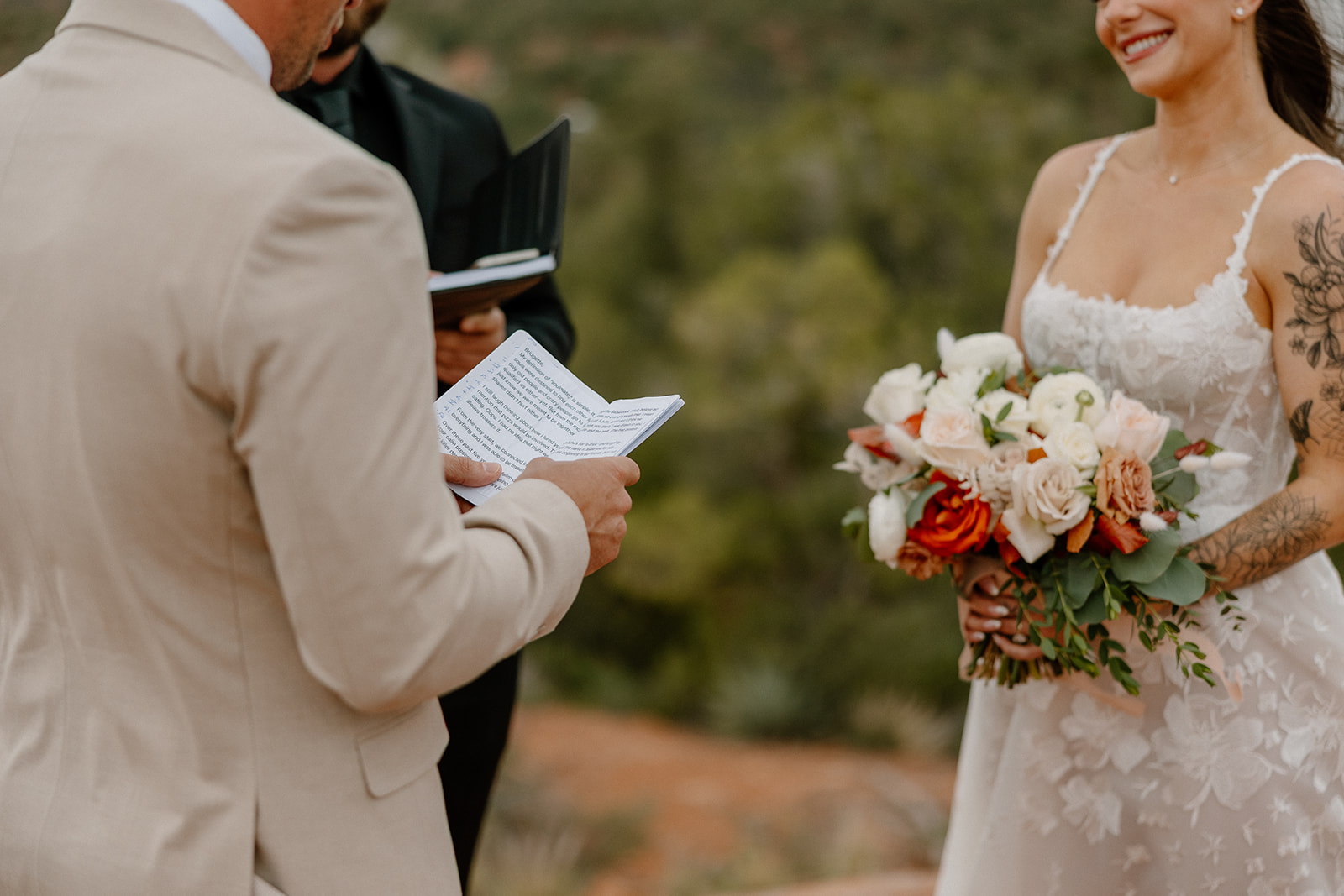 Close-up of the groom reading handwritten vows, with the bride smiling and holding a lush bouquet of white, peach, and orange blooms.