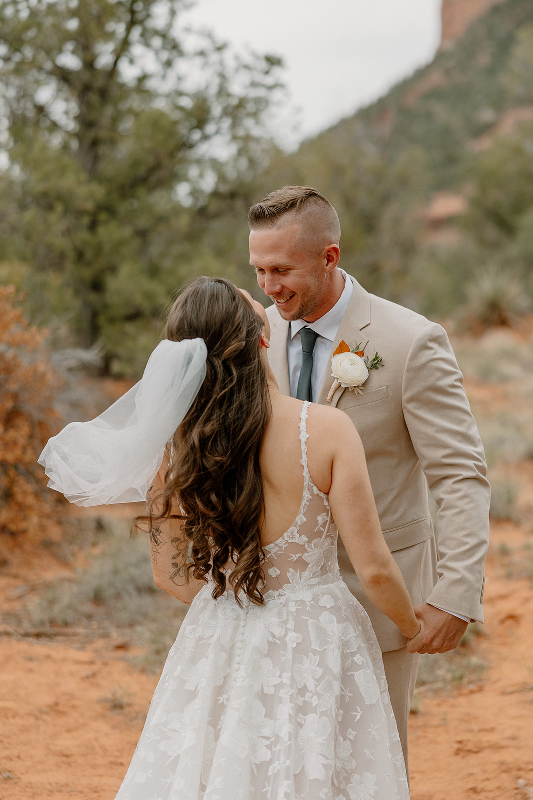 Groom smiles as he sees his bride for the first time, holding hands during their first look in the red rock desert with tall trees behind them.
