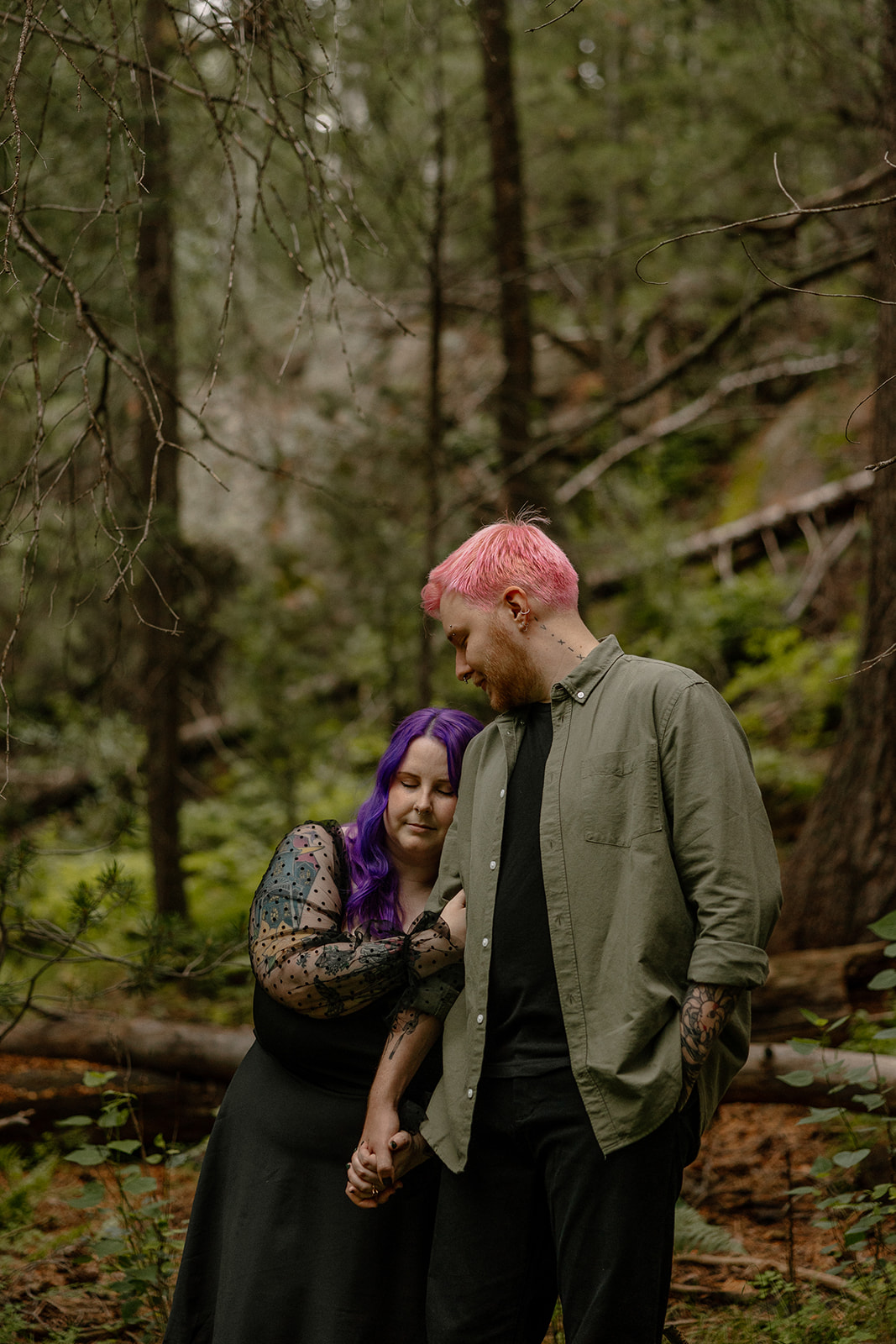 Quiet, emotional moment as a couple stands close, the woman leaning into her partner while surrounded by pine trees.