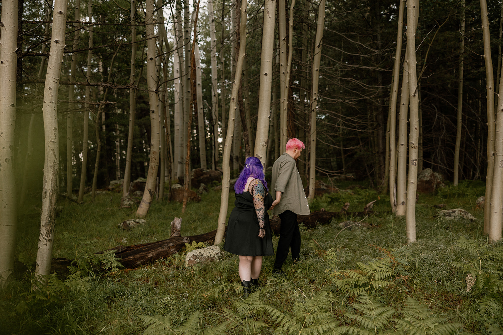 A couple walks slowly through a grove of aspen trees, holding hands as sunlight filters through the woods—natural, earthy engagement photo ideas.