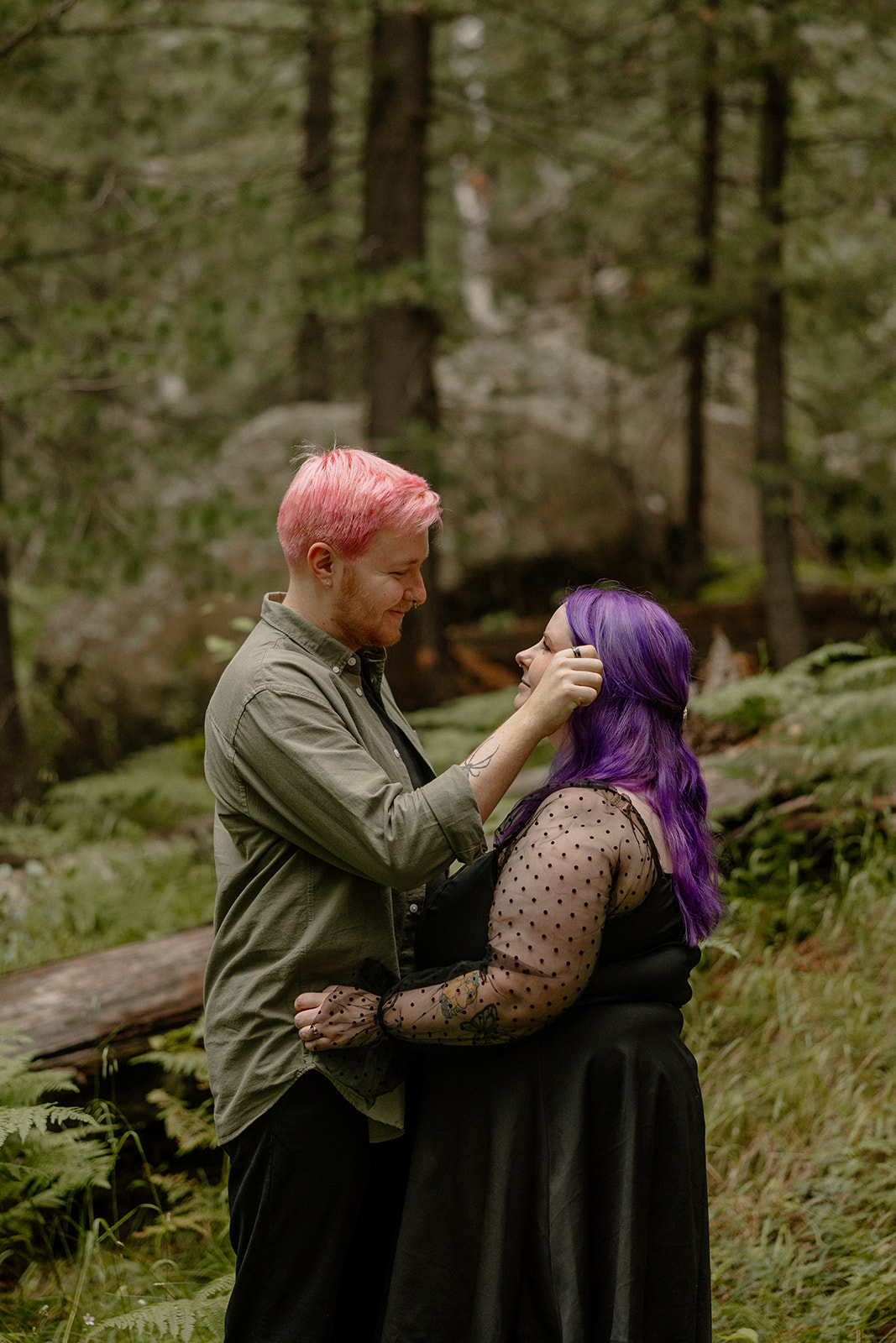 Man tucks the woman’s hair behind her ear in a gentle gesture, both framed by greenery and forest ferns.
