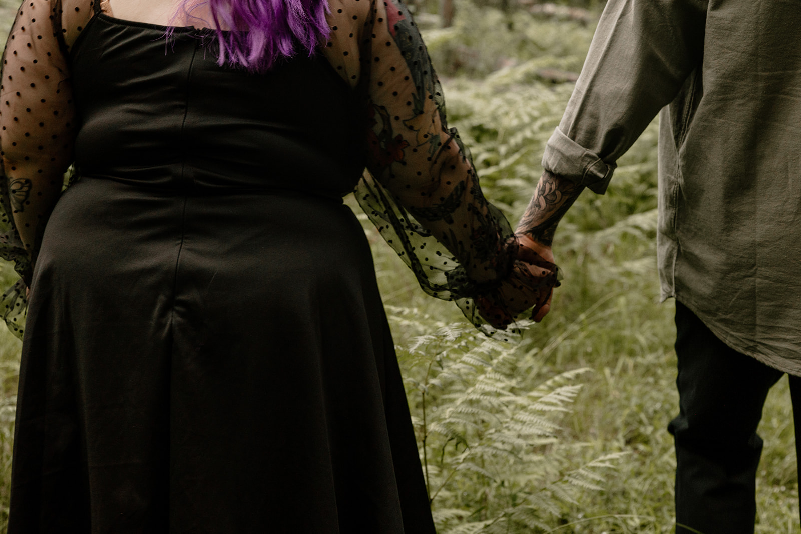 Tight crop of a couple holding hands while walking through the forest, showcasing the contrast between soft greenery and bold personal style.