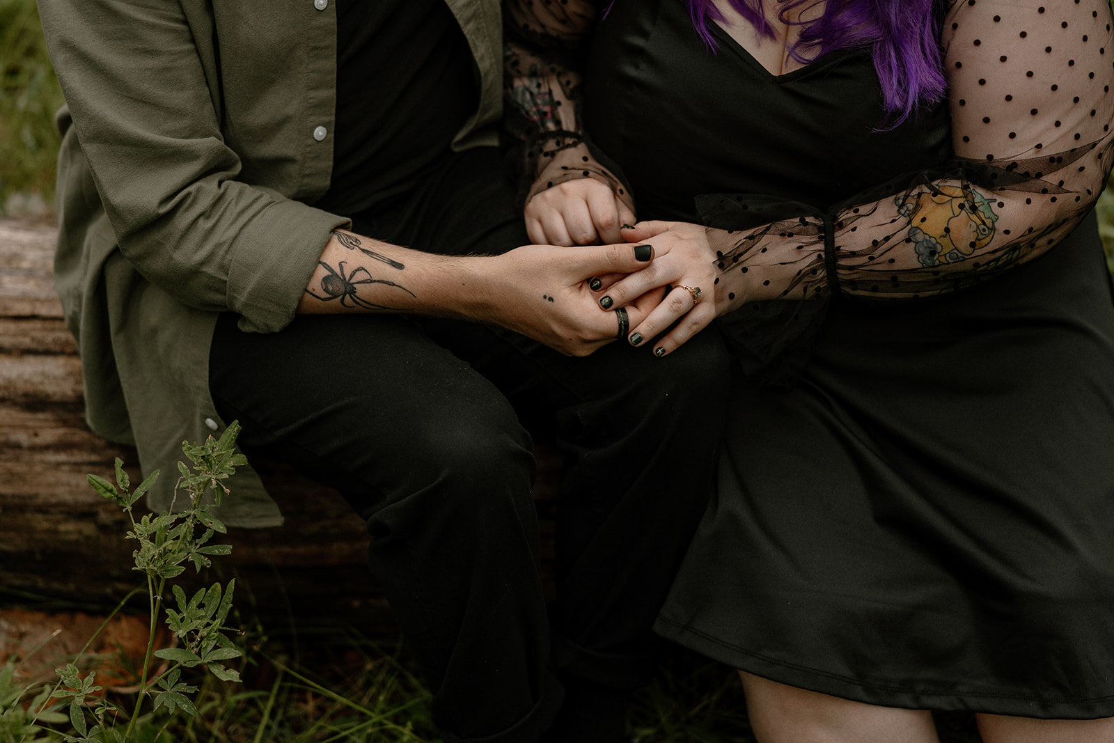 Detail shot of two people sitting close on a log, holding hands gently with tattooed arms and dark nail polish.