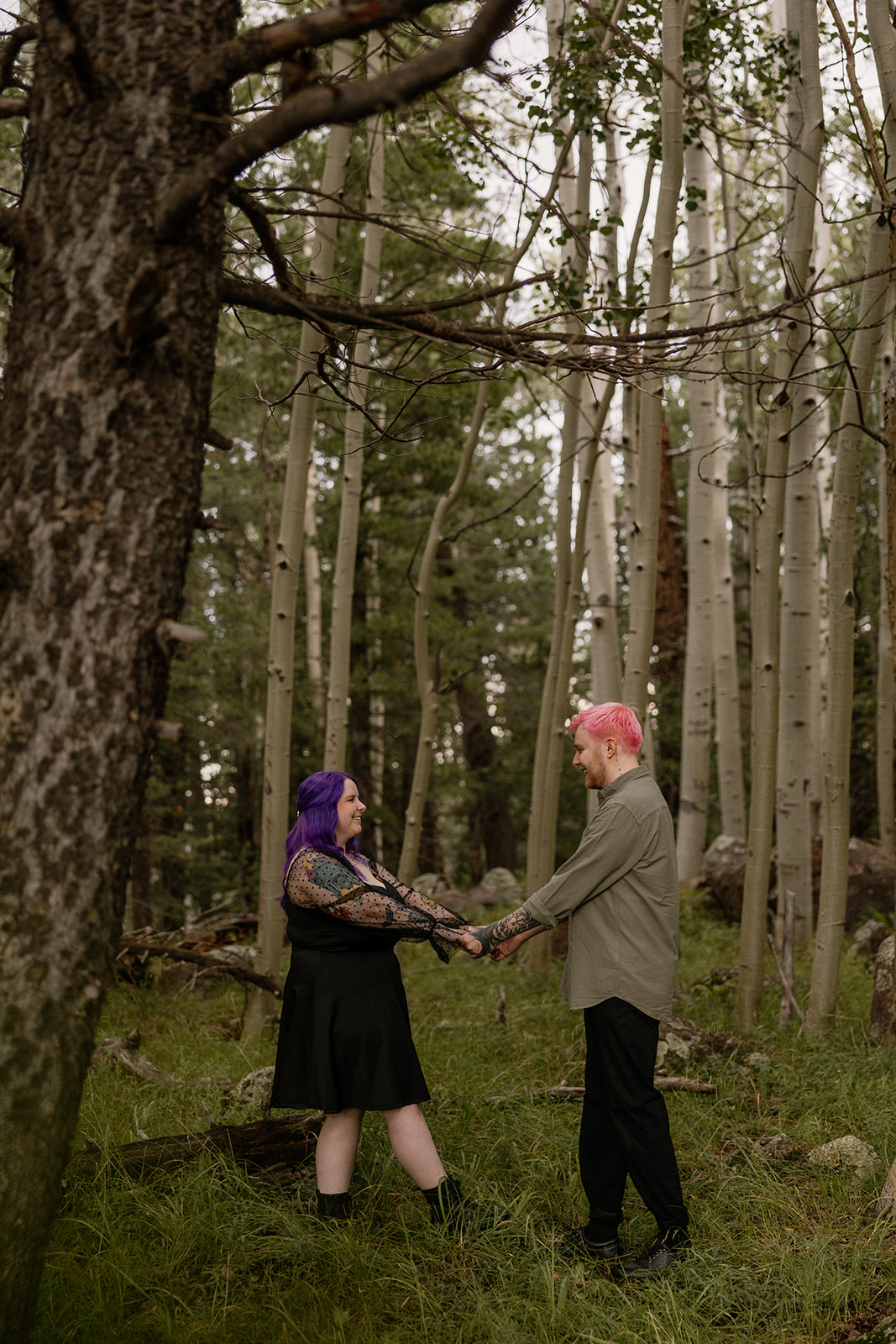 Couple standing apart but connected by clasped hands, framed by trees with a raw, woodland feel.