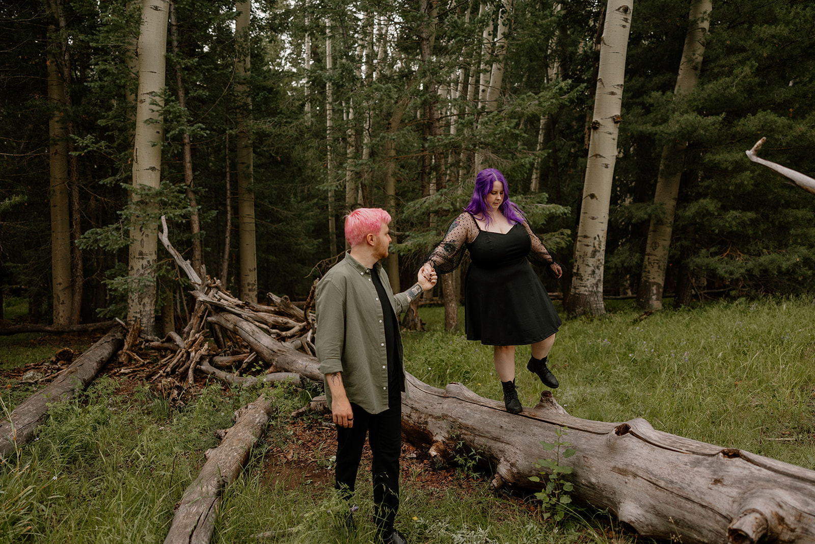 One partner balances on a fallen log while the other offers a steady hand, surrounded by tall pines and playful energy.