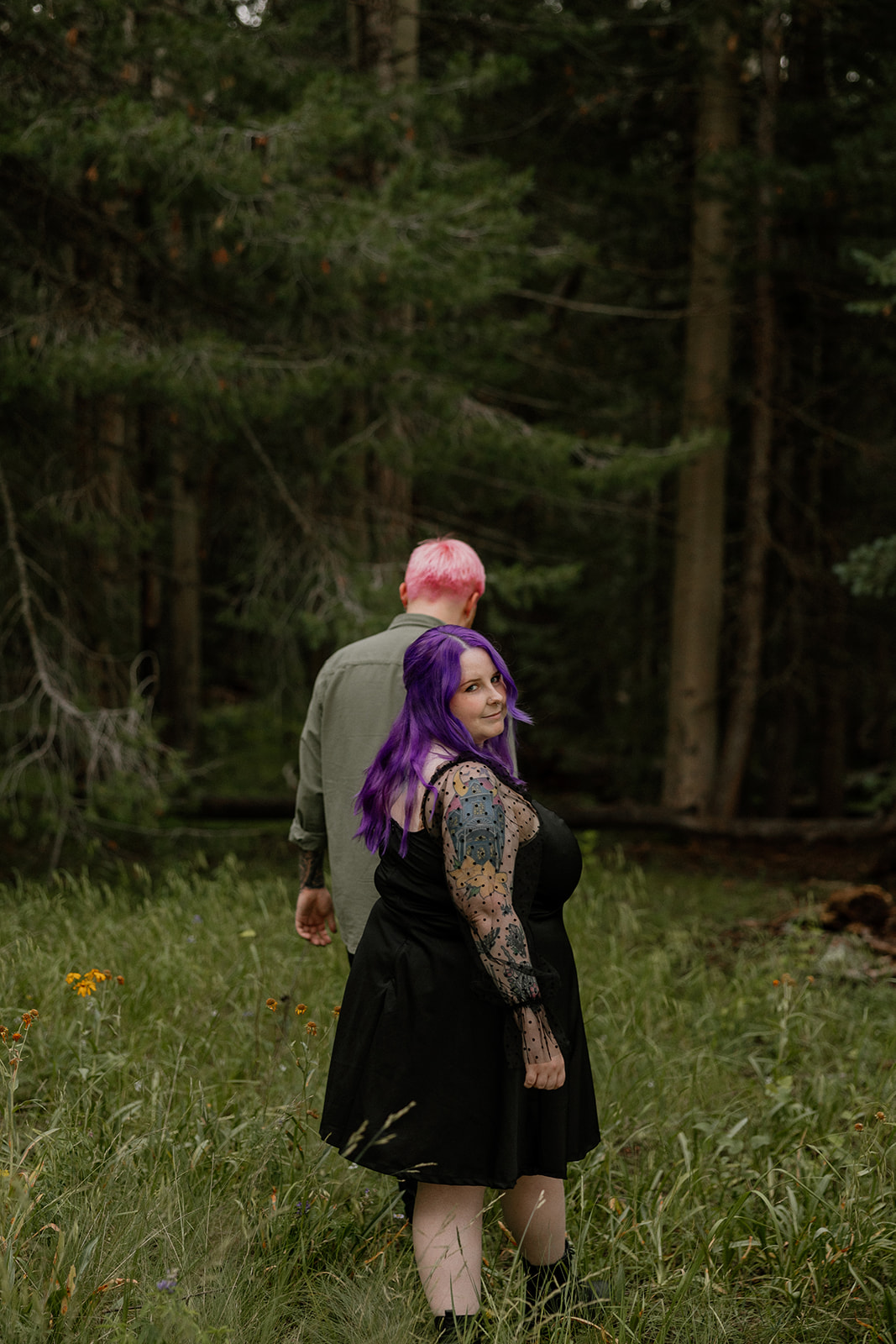 The woman looks back over her shoulder with a playful smile while walking away with her partner through tall grass and pines.