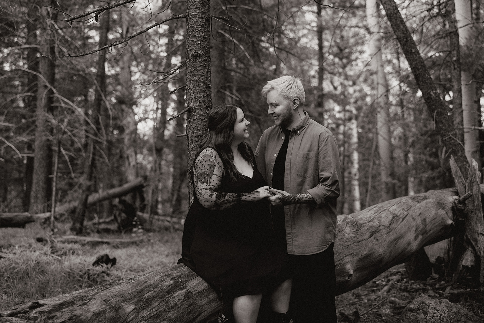 Black and white image of the couple perched on a fallen log, smiling and locked into each other’s gaze with trees surrounding them.