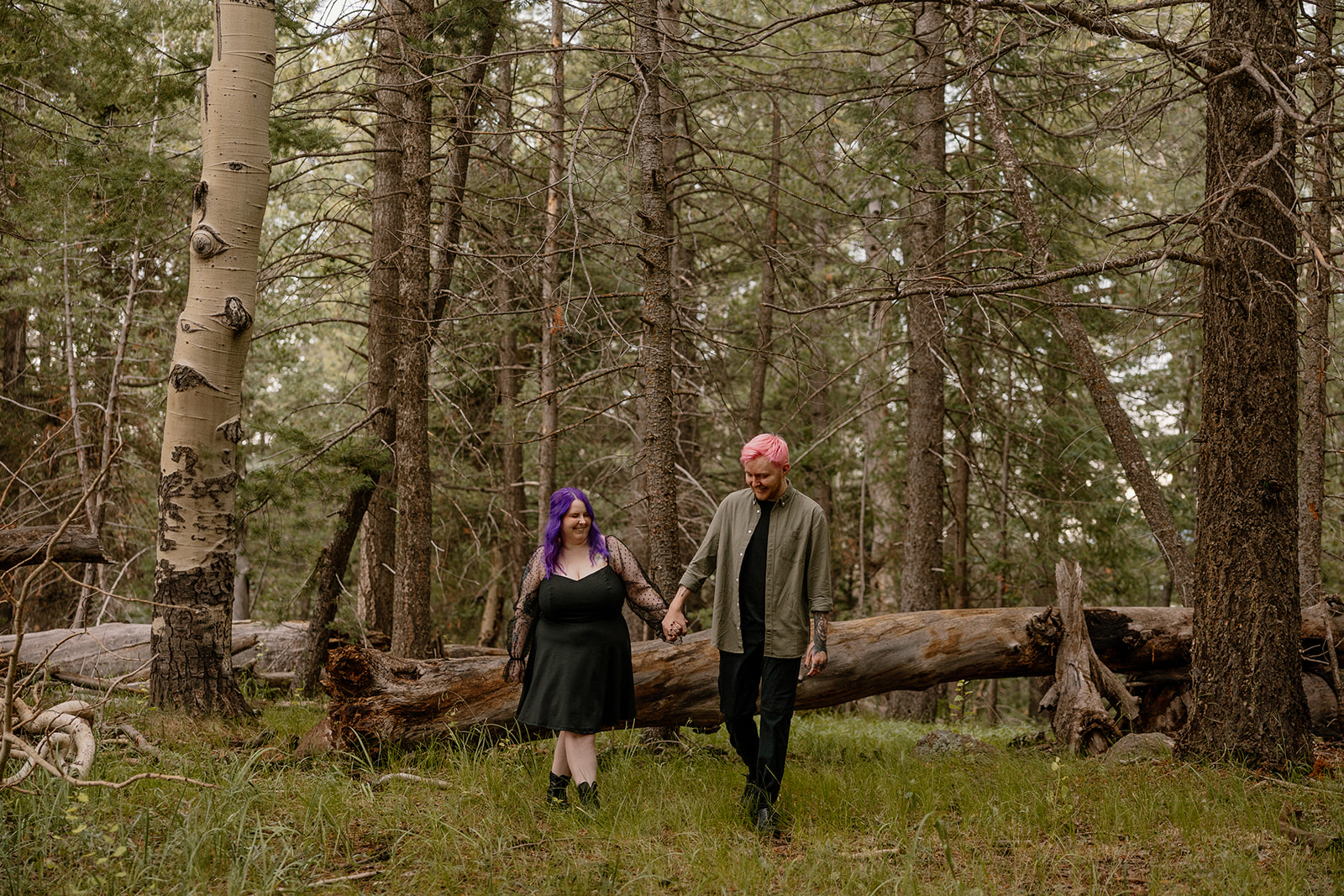 Couple walking through a clearing, smiling and holding hands in front of a fallen log deep in the woods.