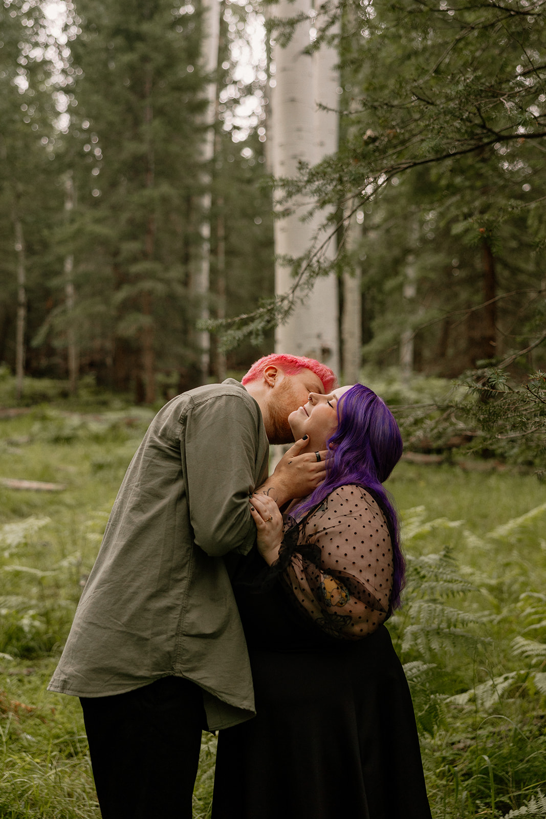 The couple shares a playful kiss, leaning into each other with joyful energy in a fern-filled forest.