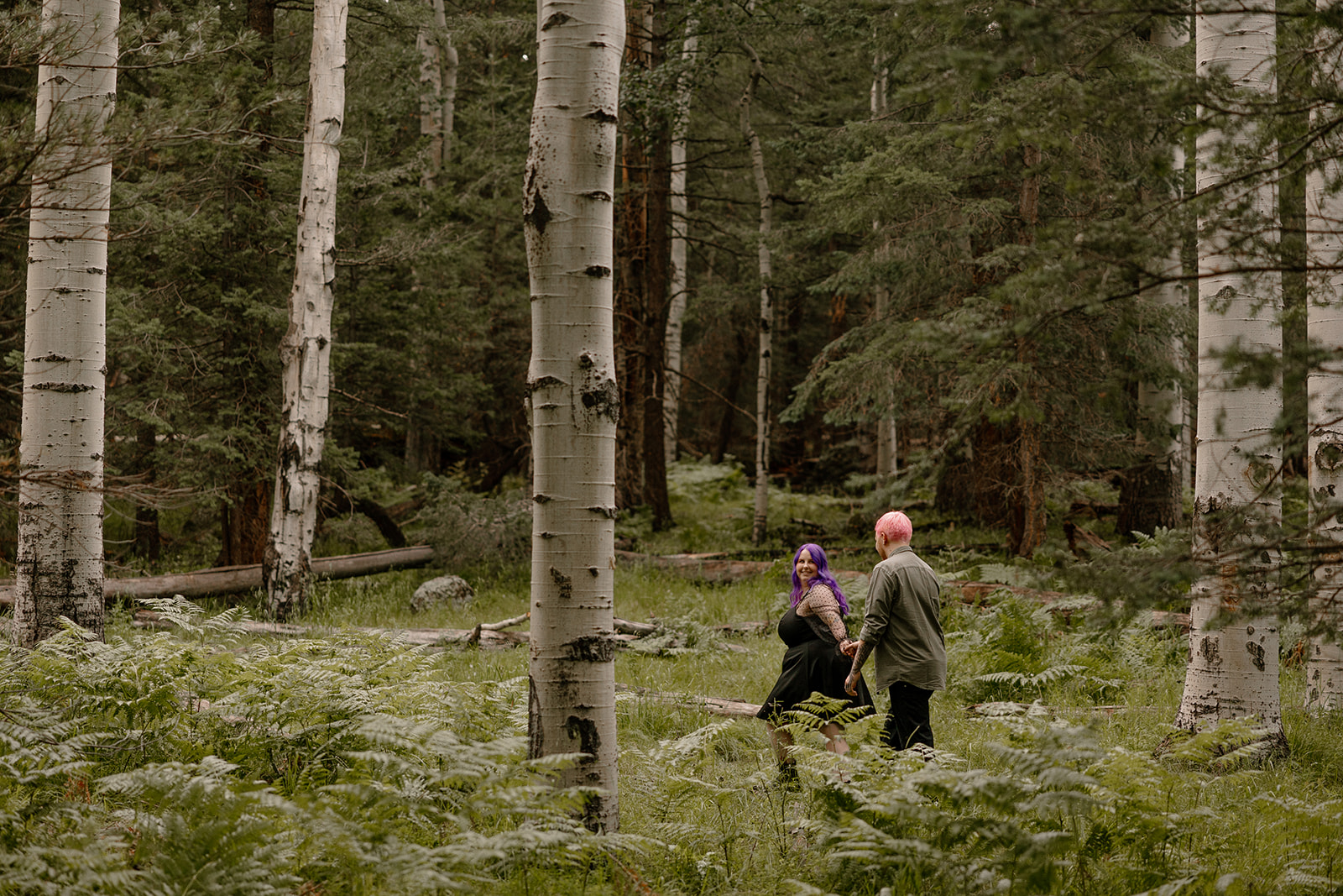 Couple walking through an aspen grove, the woman looking back with a soft smile while the man holds her hand—an editorial-style take on engagement photo ideas.