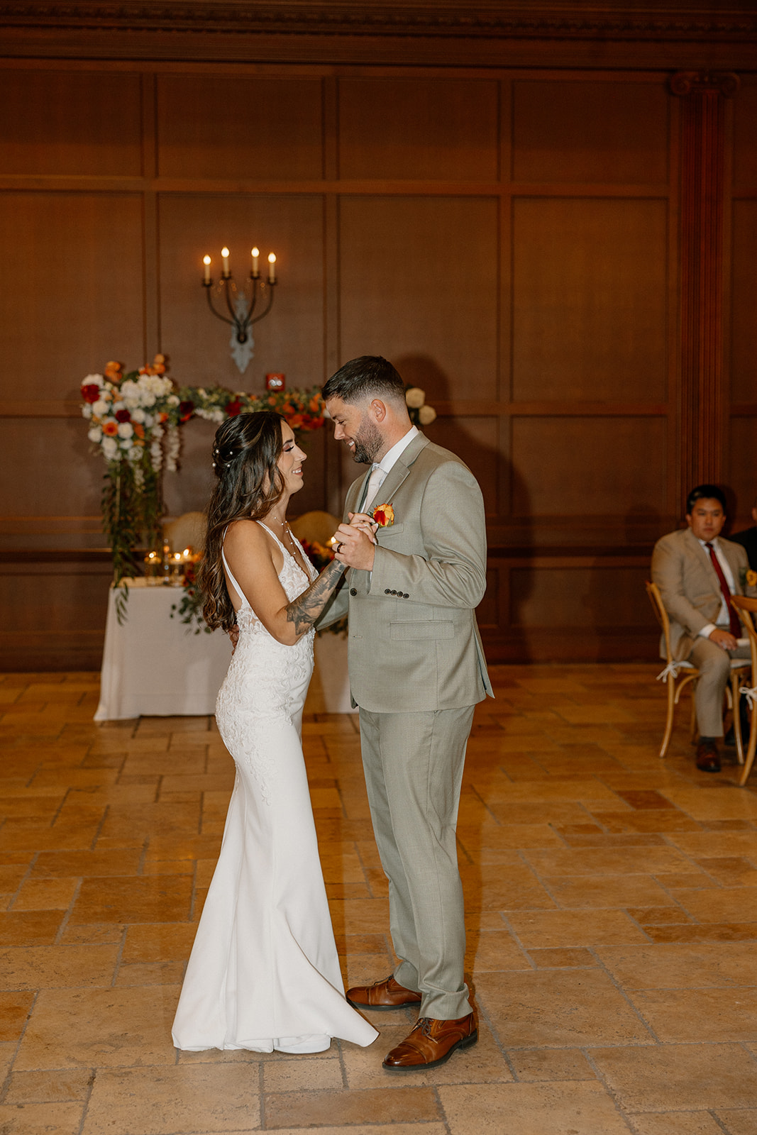 The couple gently sways together on the stone tile floor, lost in each other beneath moody lighting and classic wood-paneled walls.
