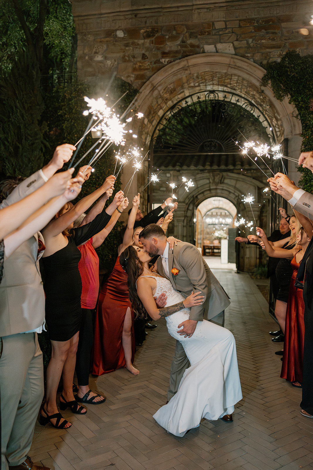 Sparkler exit moment at night as guests cheer and the couple kisses under stone archway at a Gilbert wedding venue.