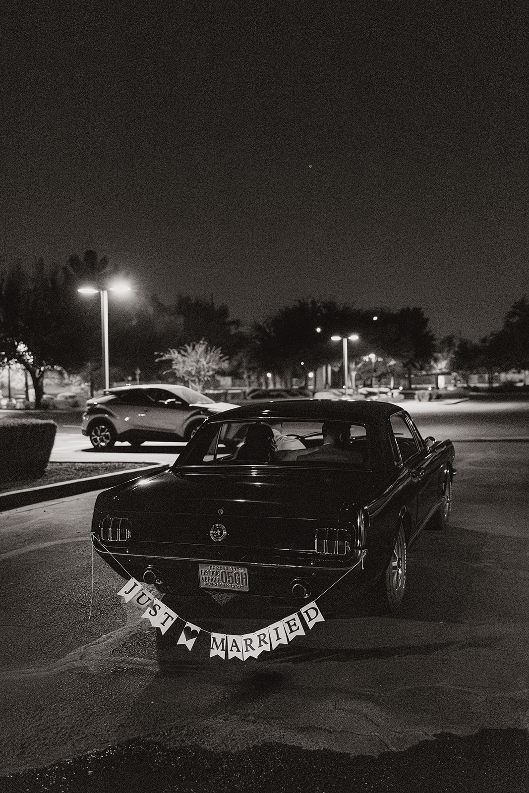Black-and-white image of a “Just Married” sign hanging from a Mustang under the night sky, headlights softly glowing in the parking lot.