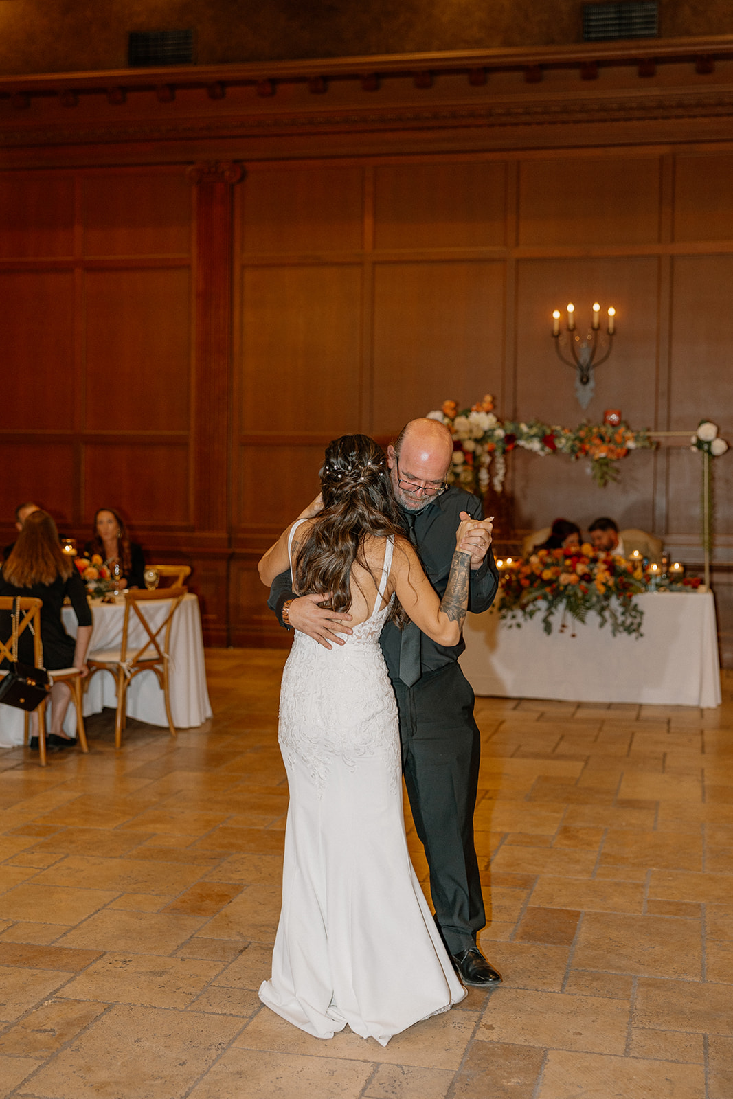 The bride dances with her father on a soft-lit reception floor, with the sweetheart table and floral arch glowing behind them.