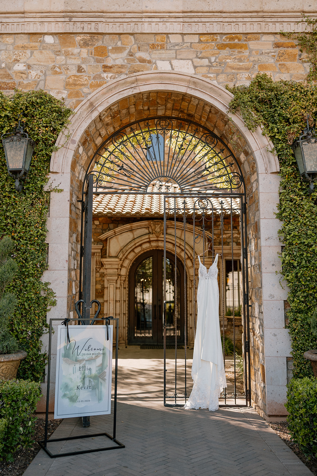 The bride’s dress hangs perfectly on a wrought iron gate beneath a stone archway framed by ivy and old-world charm.