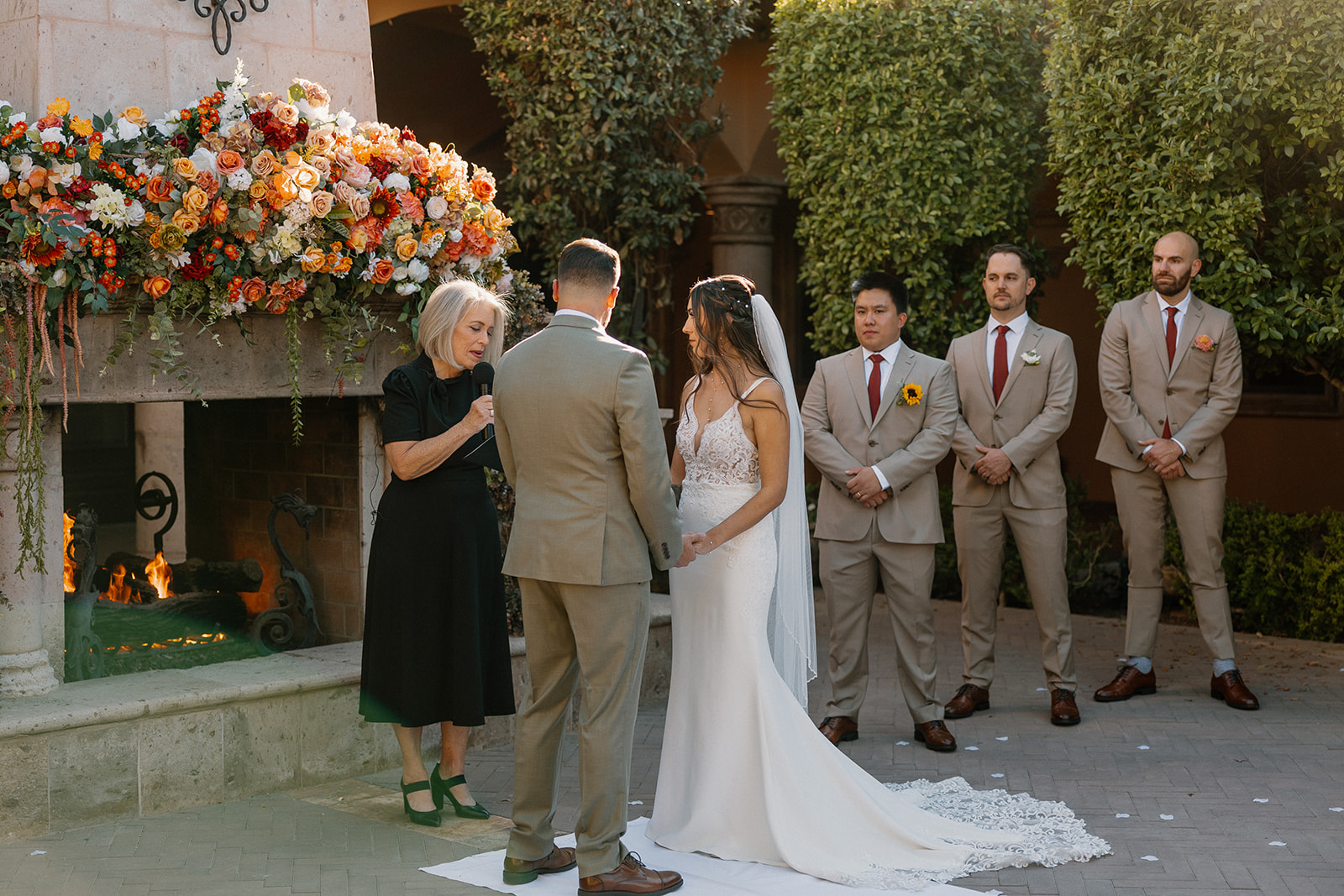 Wedding ceremony in progress as couple stands in front of floral-covered fireplace with wedding party looking on.