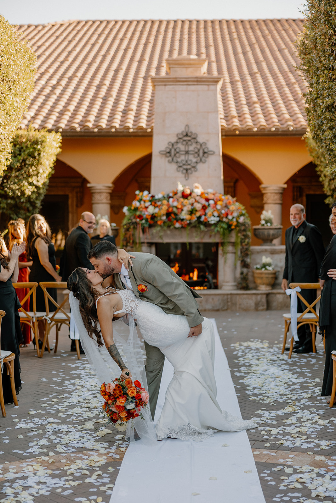 Just-married couple sharing a dramatic dip-and-kiss moment in the courtyard, surrounded by flower petals and glowing golden light.