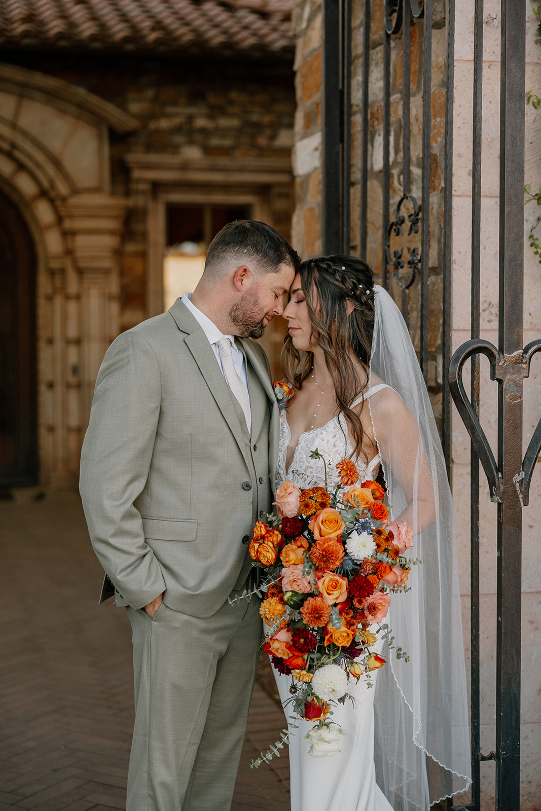 Bride and groom share a tender moment at the iron gates, framed by rustic stone walls and the bride’s vibrant bouquet of warm-toned florals.