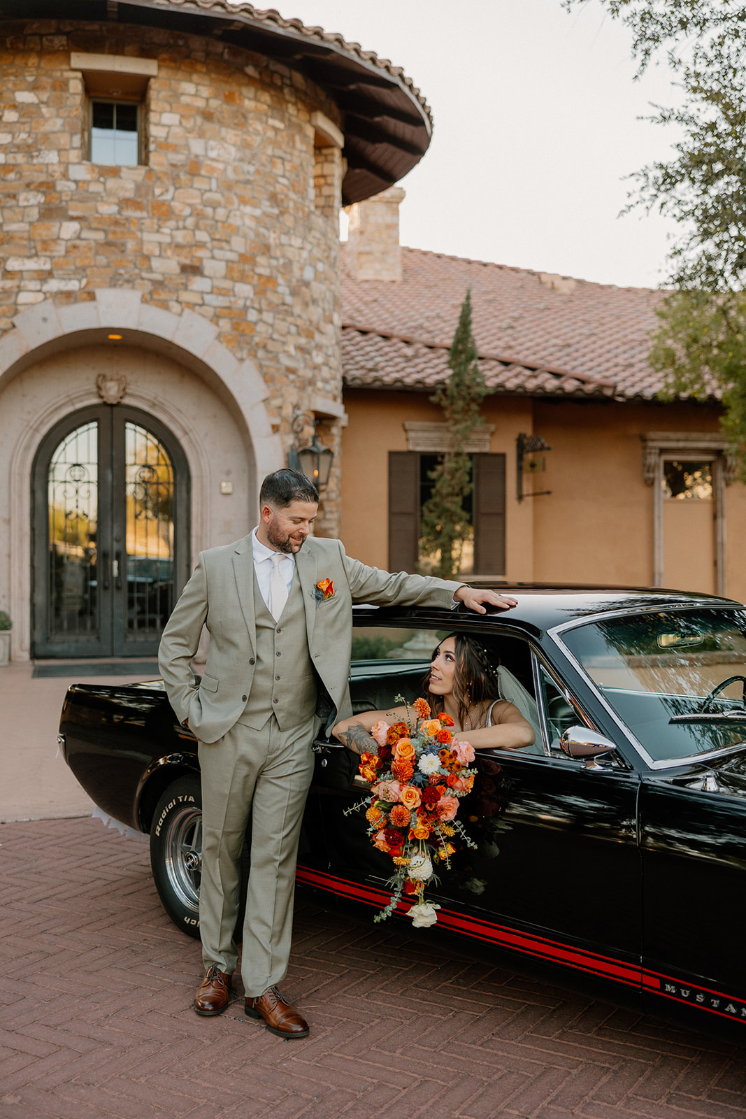 The groom leans on a black Mustang while the bride gazes up at him from the passenger seat, holding a bouquet bursting with fall tones.