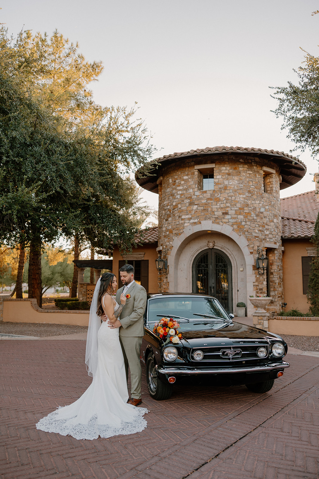 The couple shares a sweet moment beside a black Mustang, standing on the brick drive with warm evening light touching the venue’s round tower entrance.