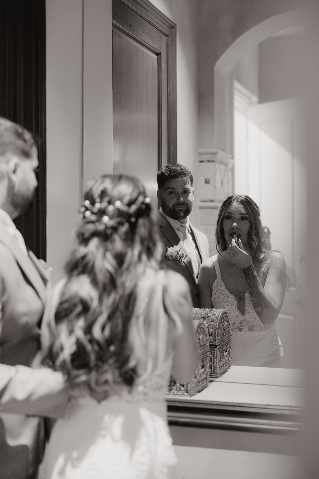 Black and white image of the bride applying lipstick as the groom watches her reflection, an intimate pause before the celebration begins.