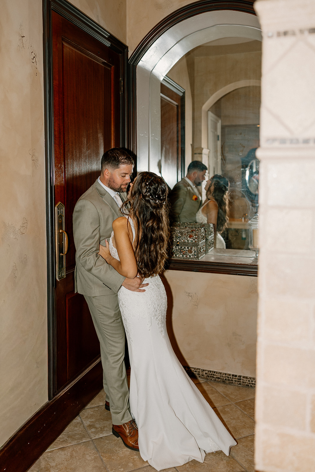 The couple shares an intimate moment by a large mirror and wooden door, tucked away from the buzz of their wedding day.