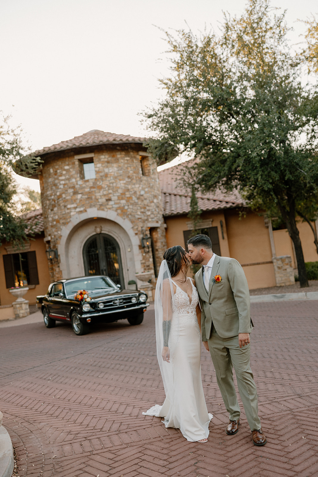 The newlyweds kiss in front of a classic black Mustang with soft evening light casting shadows on the villa-style building behind them.