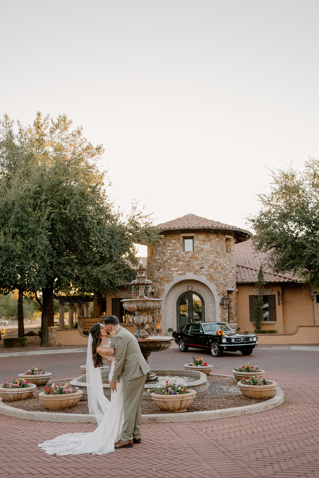 The couple shares a kiss on the brick drive near a stone fountain and villa-style entrance, with soft evening light falling over the scene.