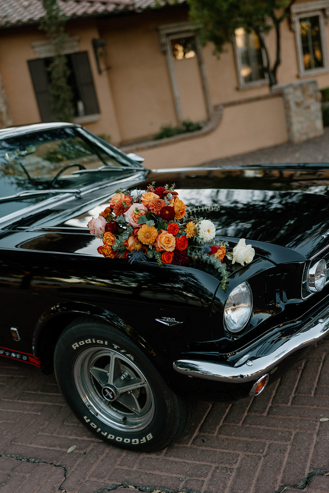 A black classic Mustang adorned with bold orange, red, and ivory flowers, parked in front of a warm-toned stucco building at a Gilbert wedding venue.