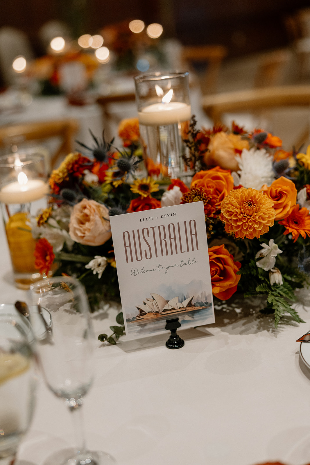 Travel-inspired wedding centerpiece featuring orange and white florals, candles, and an “Australia” table card.