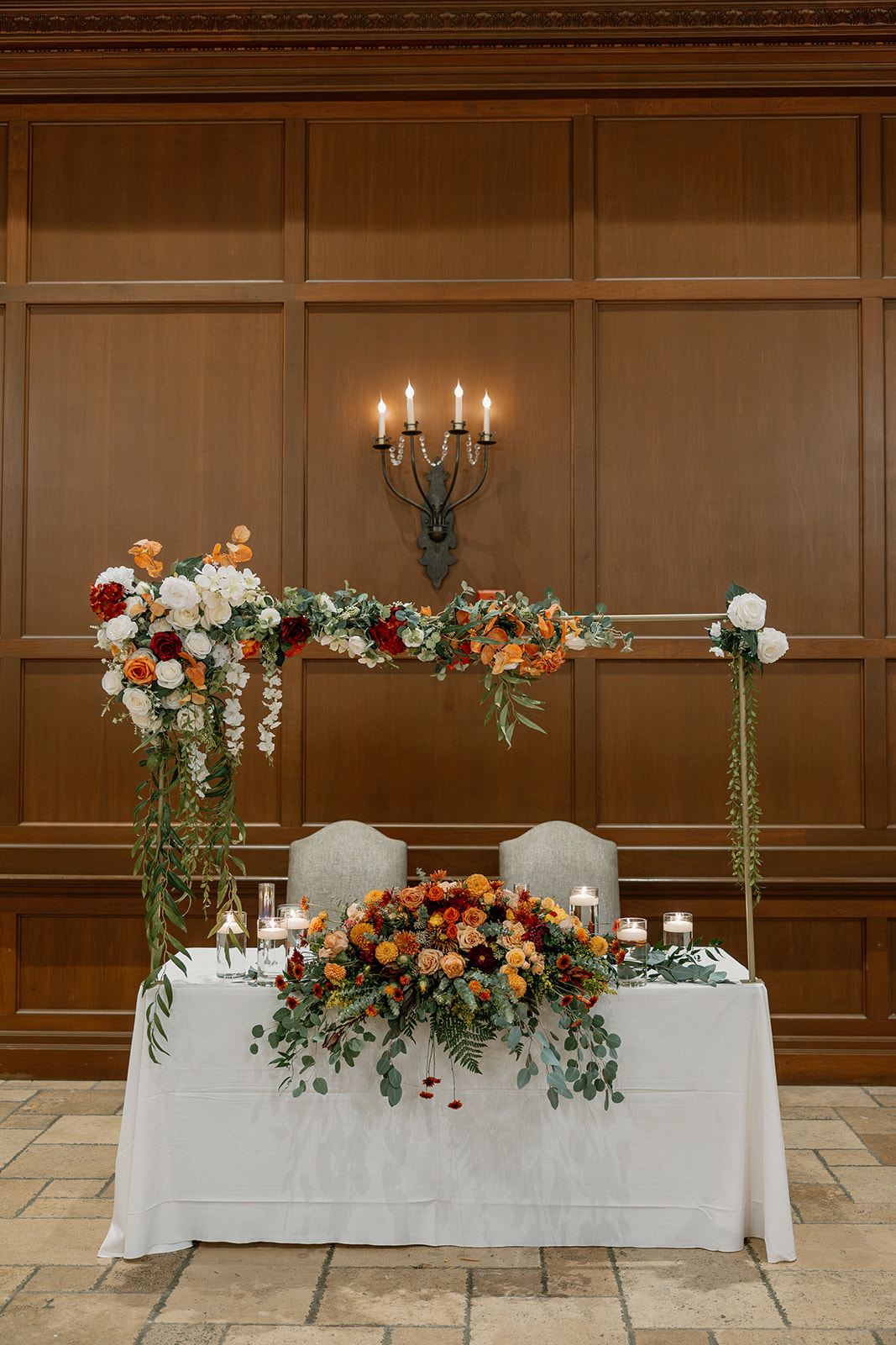 A romantic sweetheart table styled with cascading greenery, candles, and vibrant blooms, set against a wood-paneled wall and vintage chandelier.