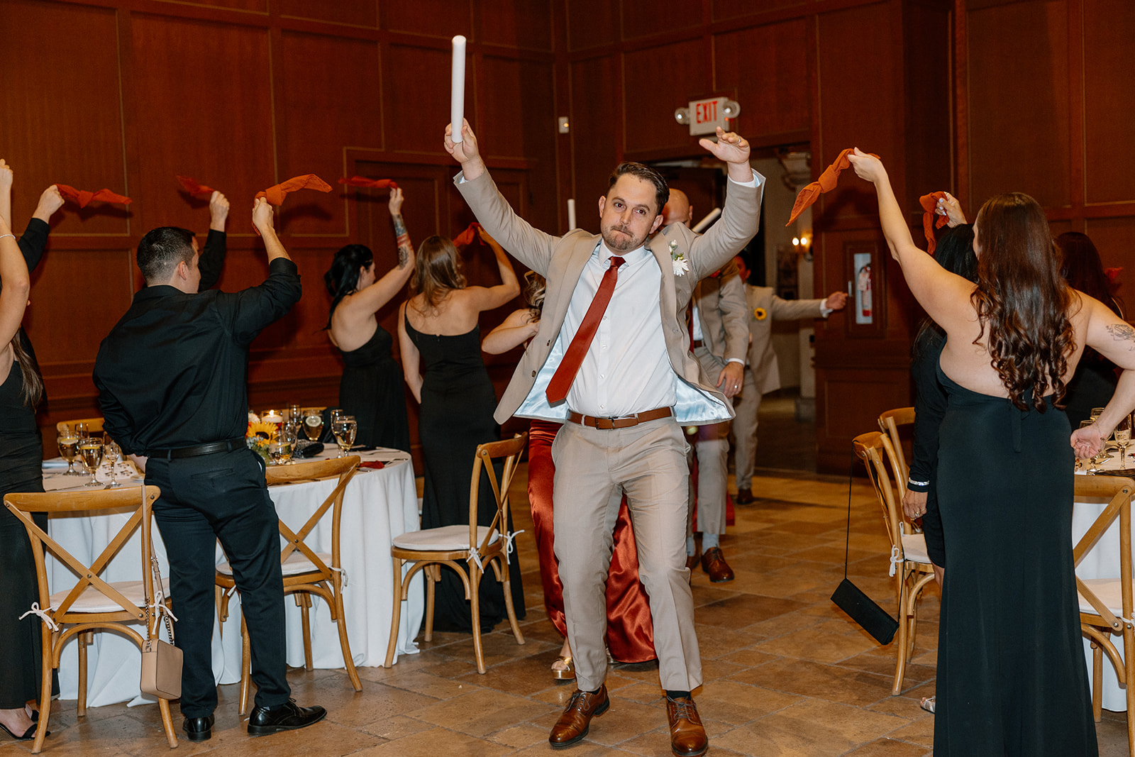 A groomsman dances into the reception waving a glow stick, hyping up guests lined up with ribbons and cheers at a Gilbert wedding venue.