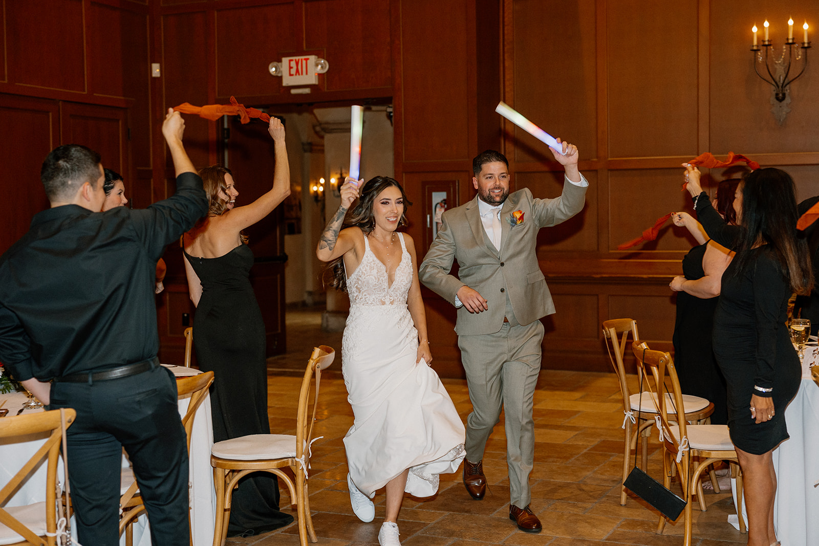 Newlyweds dance their way into the reception, surrounded by cheering guests waving glow sticks and ribbons inside a warmly lit ballroom.