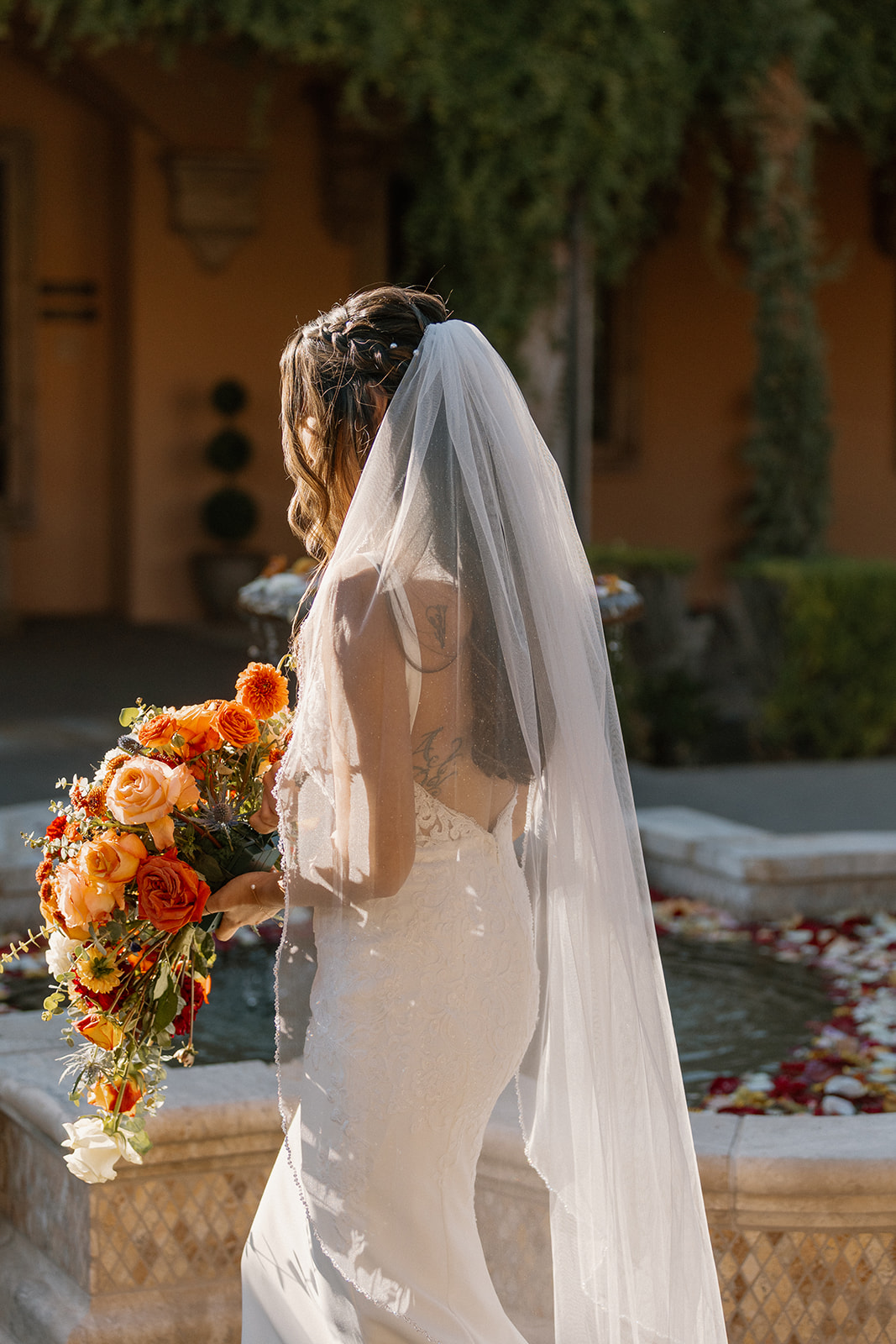 The bride holds her bouquet while sunlight glows through her veil, casting a dreamy silhouette in front of the floral-filled fountain.
