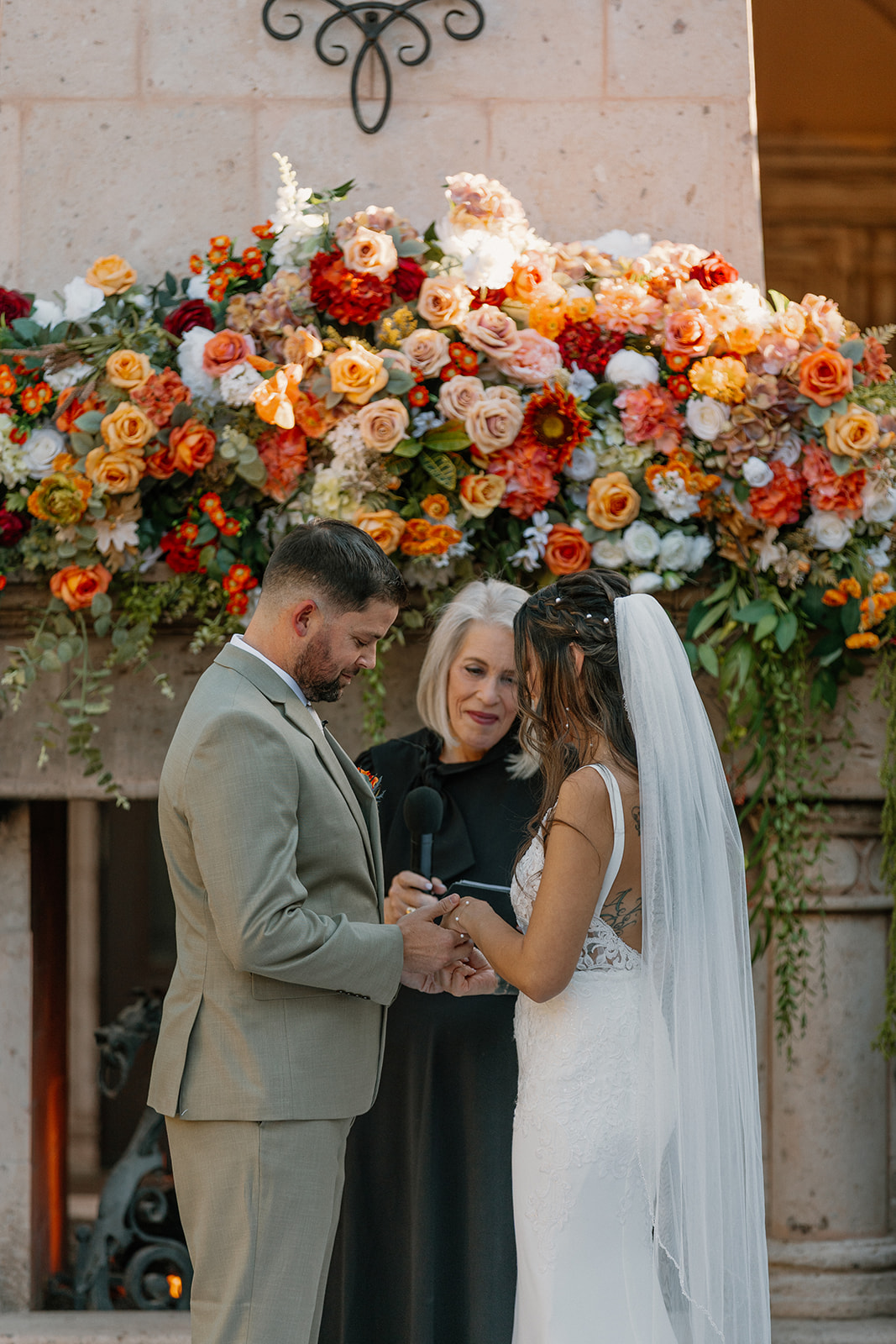 Bride and groom exchange vows with a vibrant wall of florals behind them, set against a stone fireplace that anchors the courtyard ceremony at a Gilbert wedding venue.