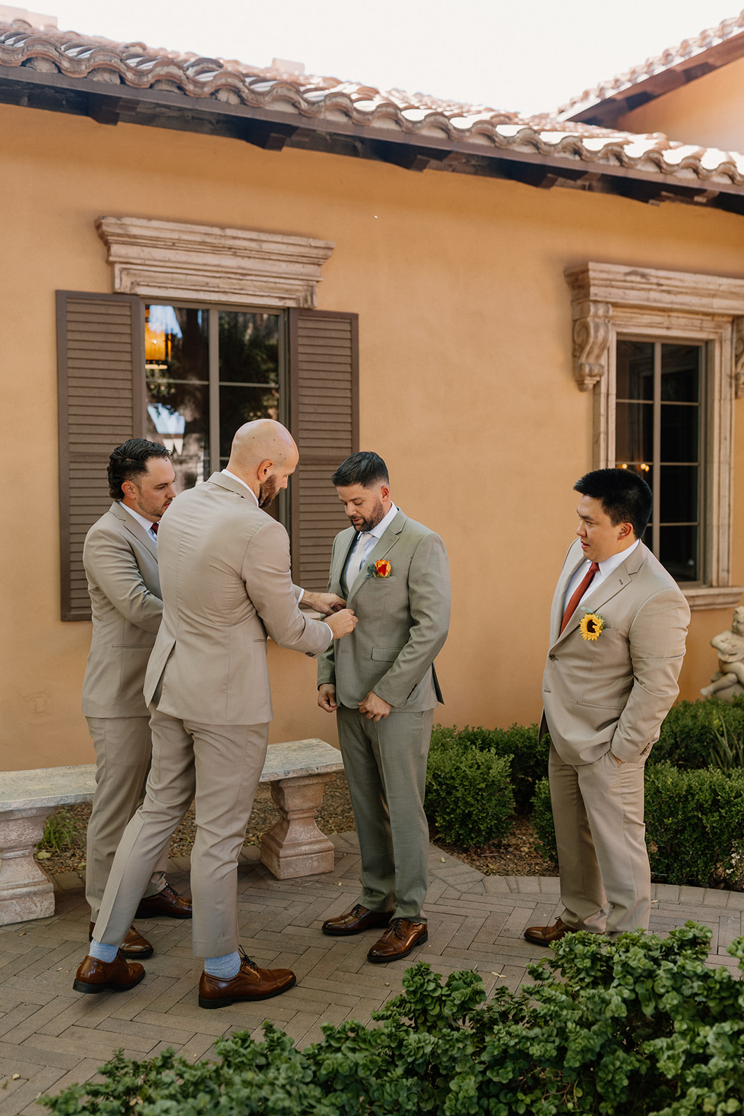 The groom gets buttoned up while his guys gather around, surrounded by neutral-toned walls, greenery, and morning light.