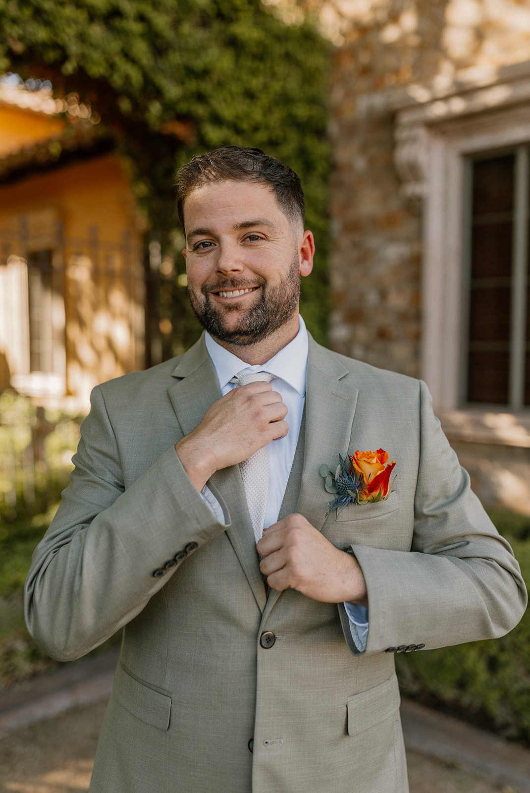 A close-up of the groom smiling and fixing his tie, boutonniere perfectly in place, framed by ivy-covered walls and golden sunshine.