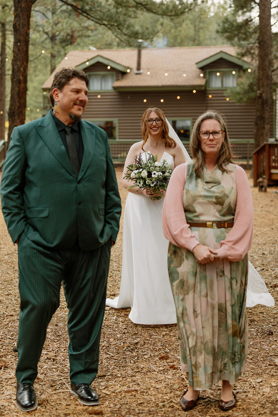 Bride smiles in the background as her parents prepare to walk her down the aisle, the cabin twinkling behind them at Nordic Village AZ.