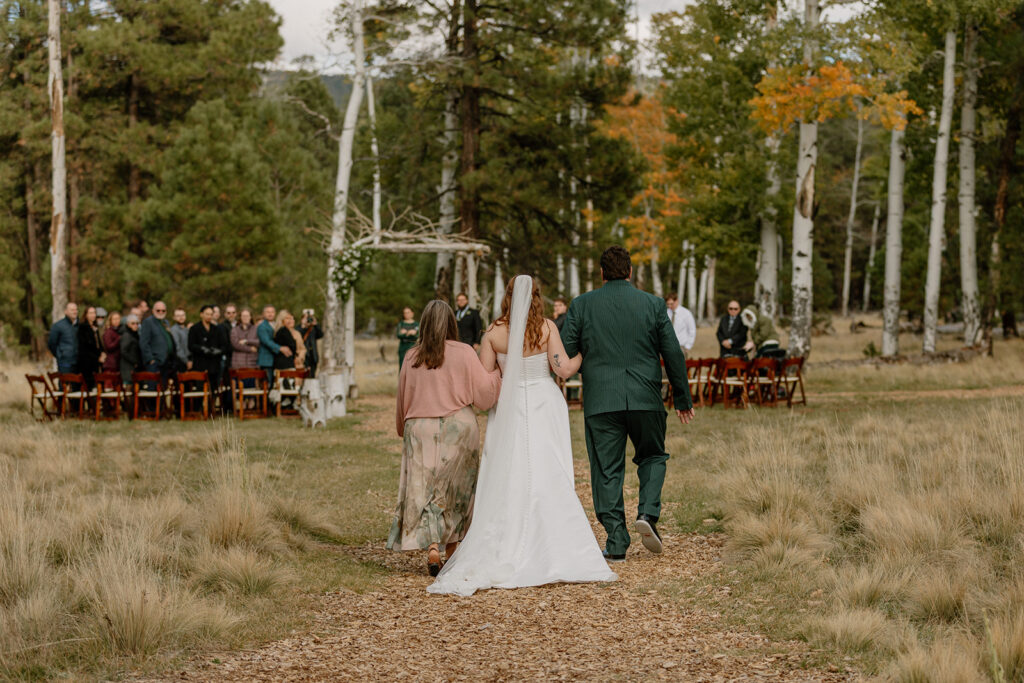 Bride walks hand-in-hand with her parents down a wooded aisle lined with tall grass, leading to a birch arch at Nordic Village AZ.