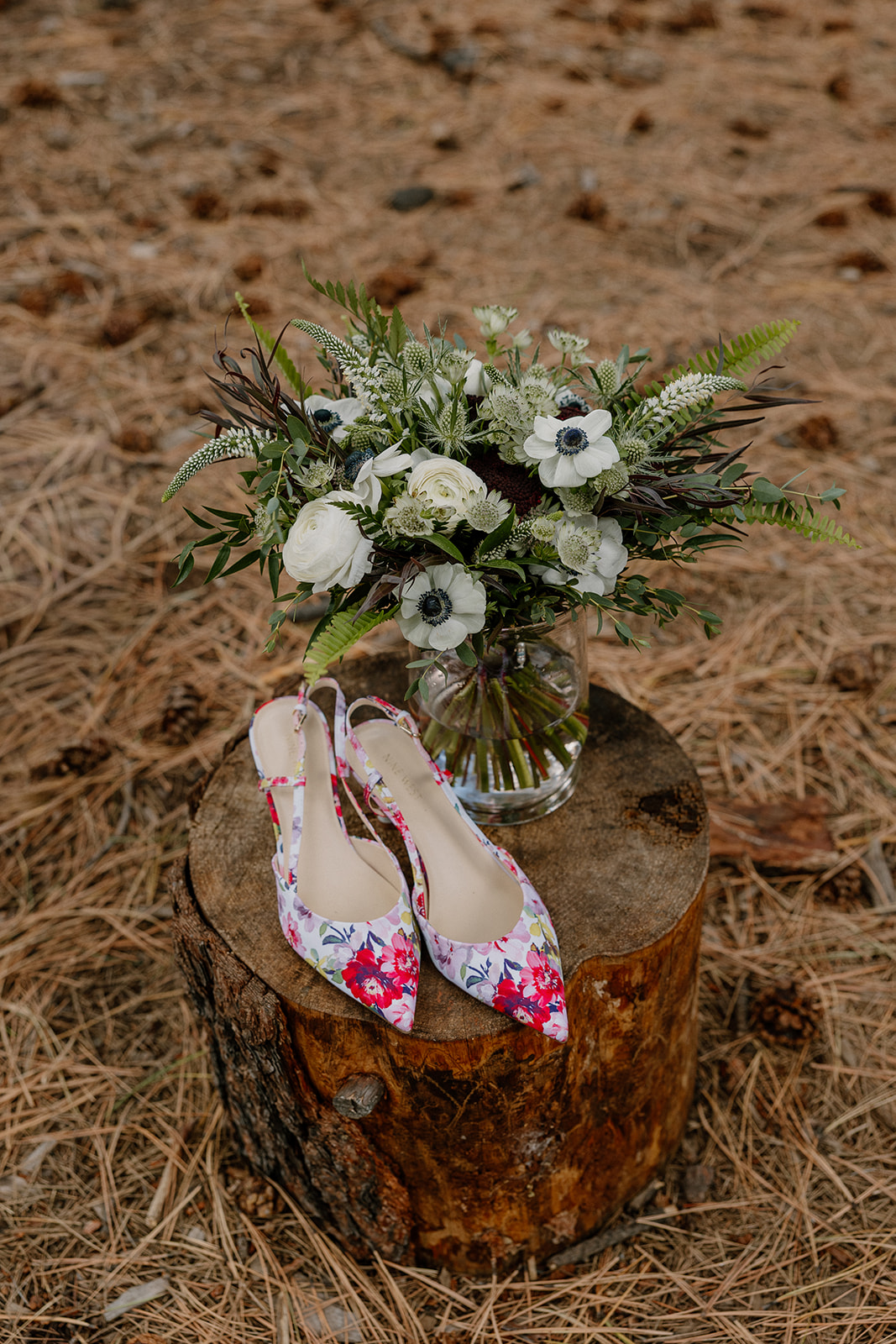 Bright floral heels and a white-and-green bouquet styled on a tree stump, capturing playful wedding details in the Nordic Village AZ forest setting.