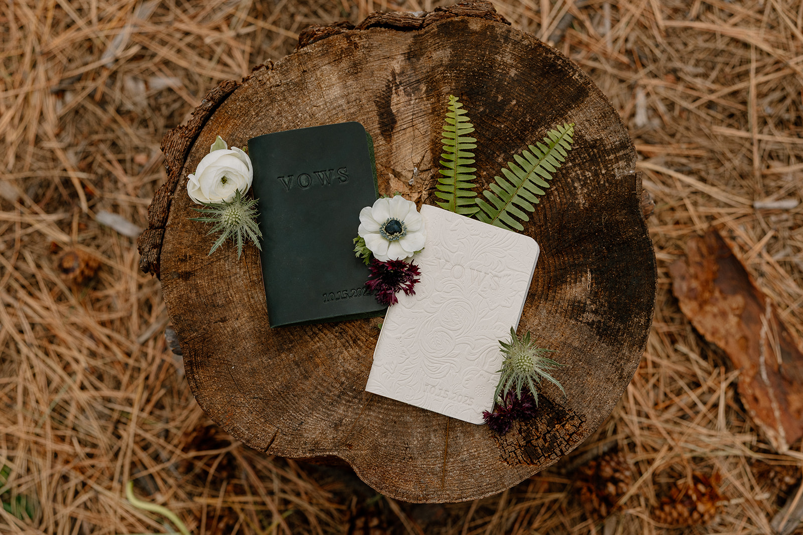 Flat lay of his and hers vow books with fresh florals and ferns, styled on a tree stump surrounded by pine needles at Nordic Village AZ.