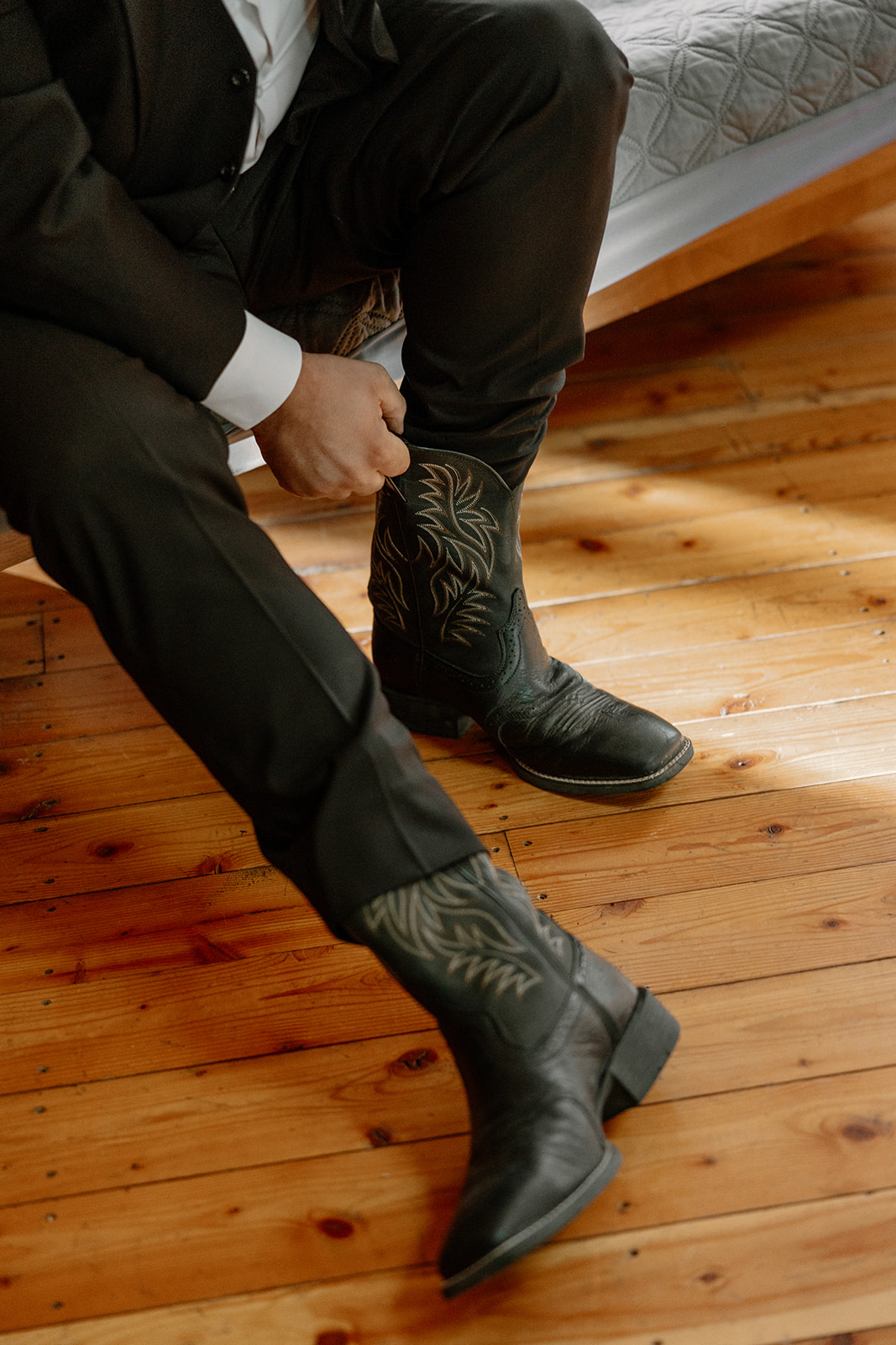 Close-up of groom slipping on embroidered black cowboy boots, captured on a rustic wood floor.