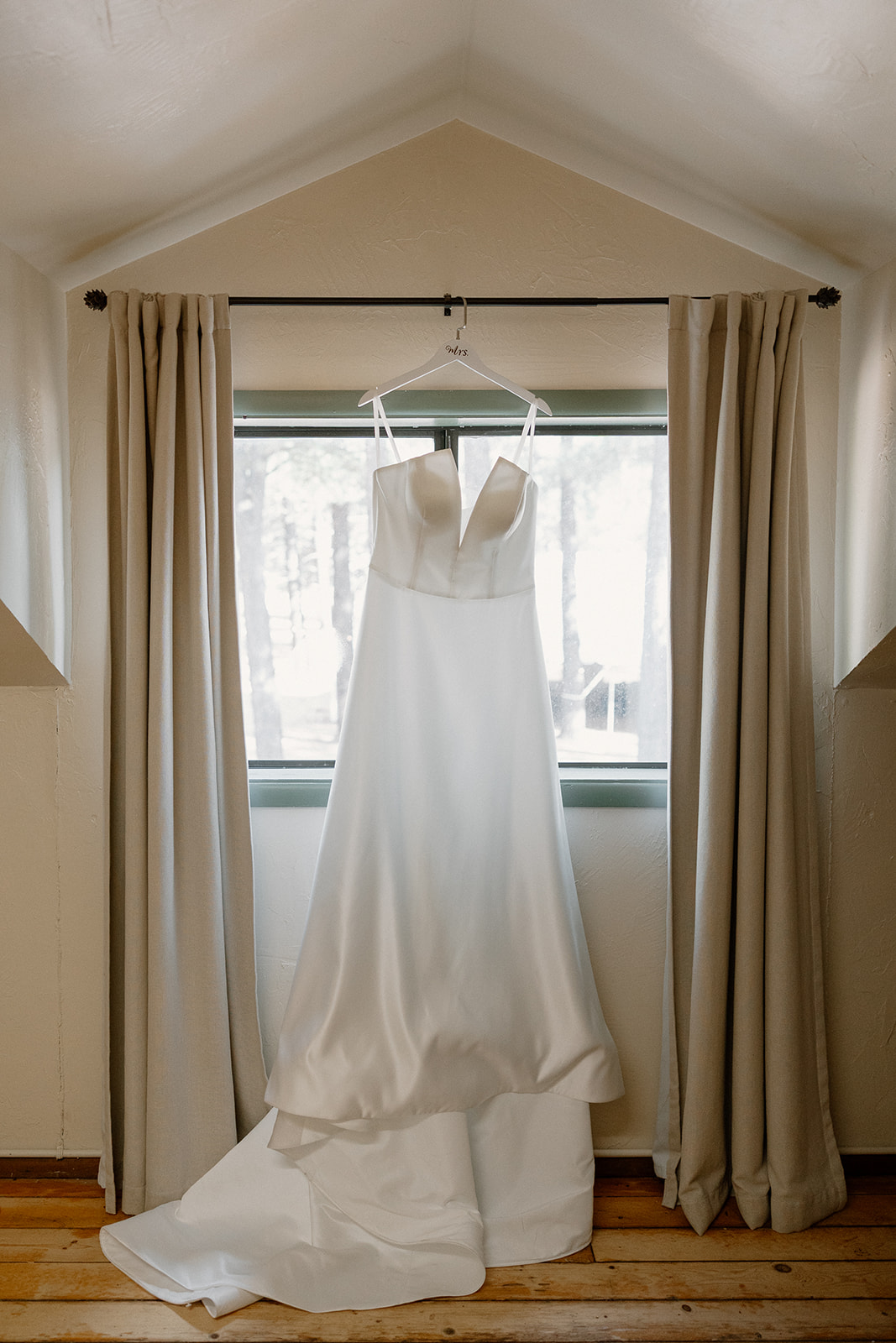 Elegant white wedding dress hanging in a cozy cabin window, soft light falling across the wood floors.