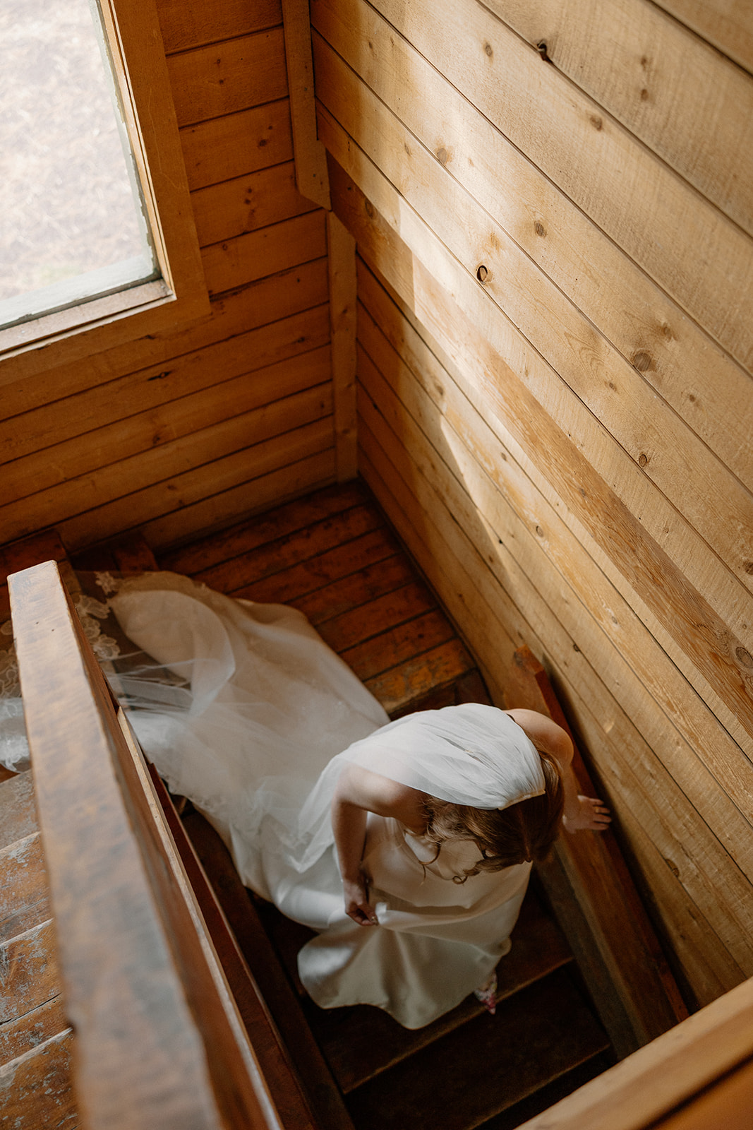 Bride gazes out the upstairs window of a woodsy cabin, her veil softly catching the light from behind the glass.