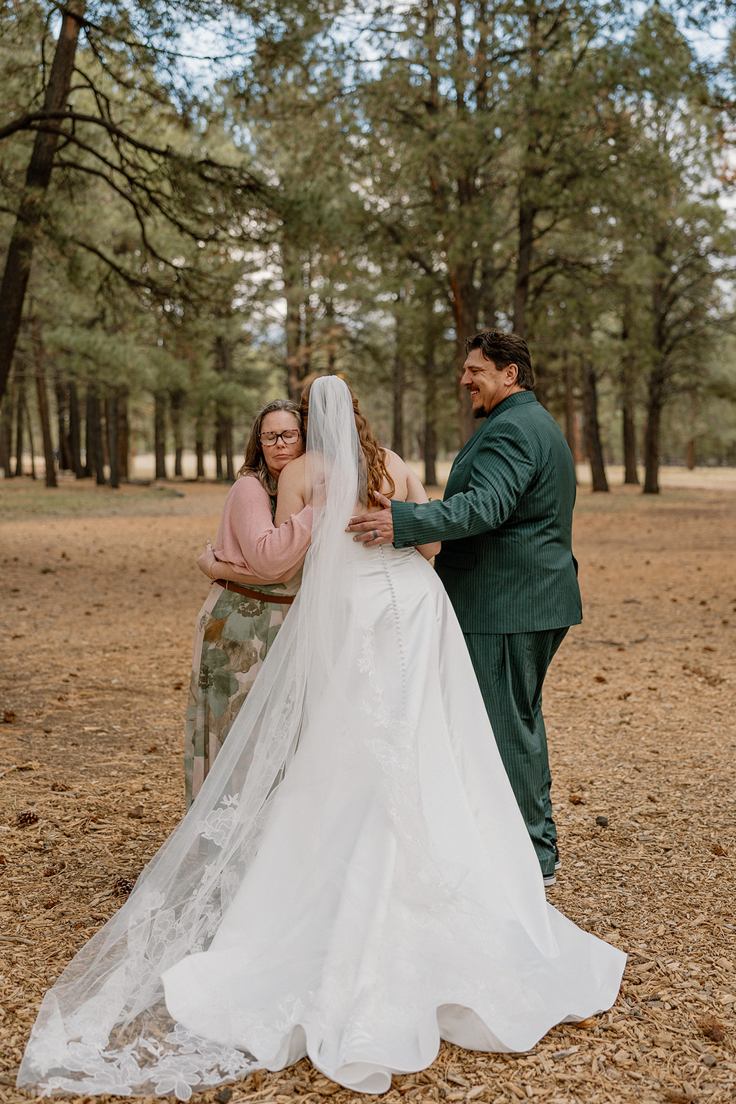 The bride approaches her parents for the walk down the aisle, surrounded by towering pine trees and soft forest light.